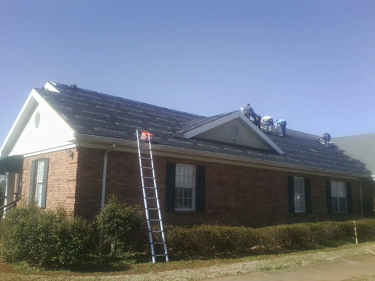 Crew tearing shingles off a brick ranch roof with a ladder set — Emergency Roof Repair work by our crew in Muenster