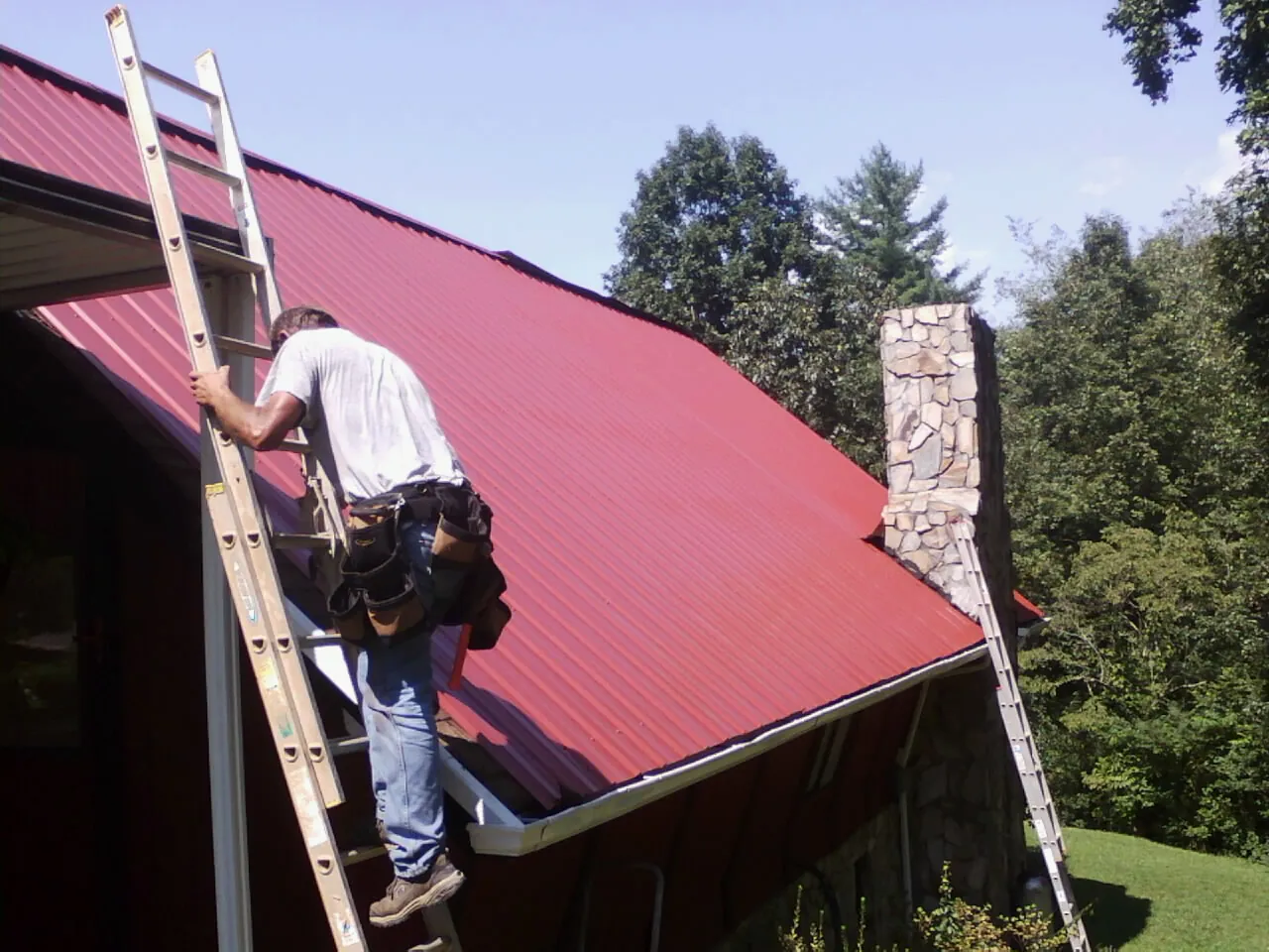 Roofer on a ladder beside a red metal roof and stone chimney — Soffit & Fascia Repair by Roofing Brothers in Muenster